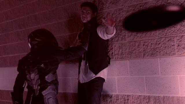 An armored figure pins a man against a brick wall as he raises his hand