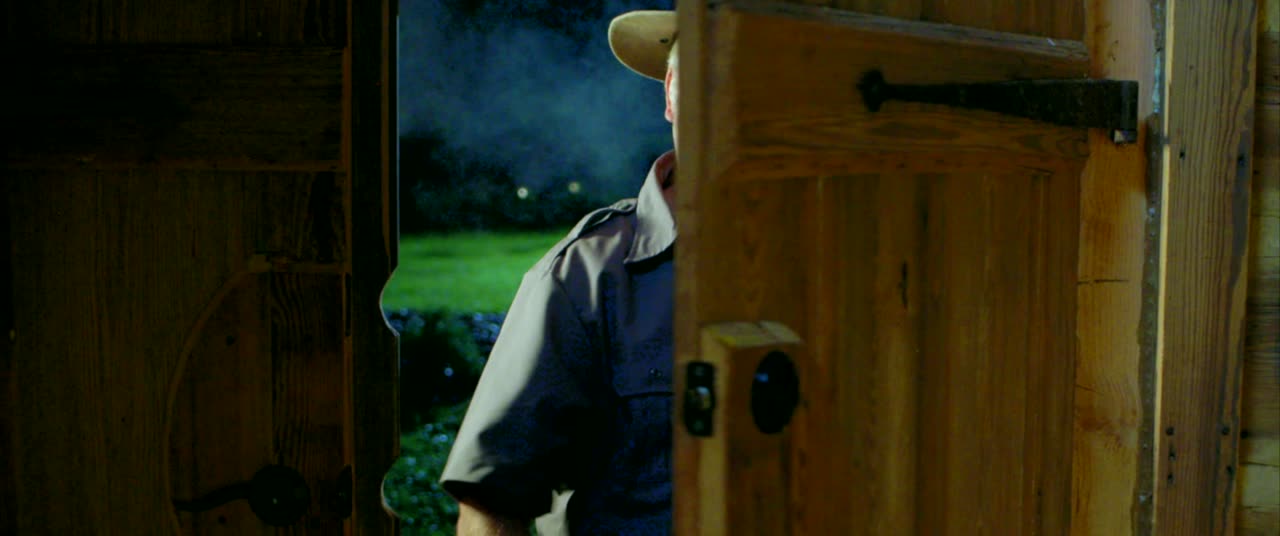 A man in a hat peers around a wooden door at night