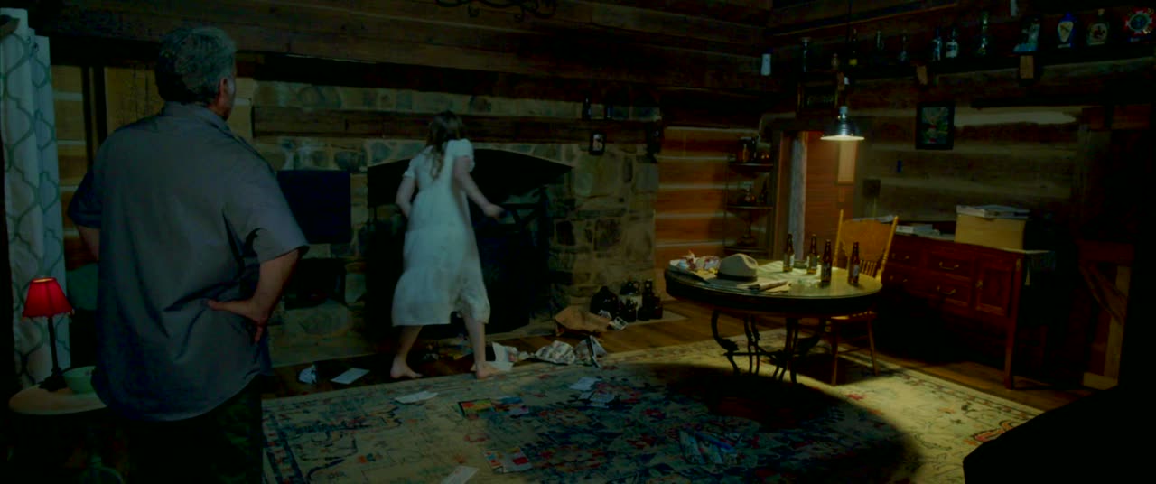 A man watches a girl in a white nightgown by a cabin fireplace